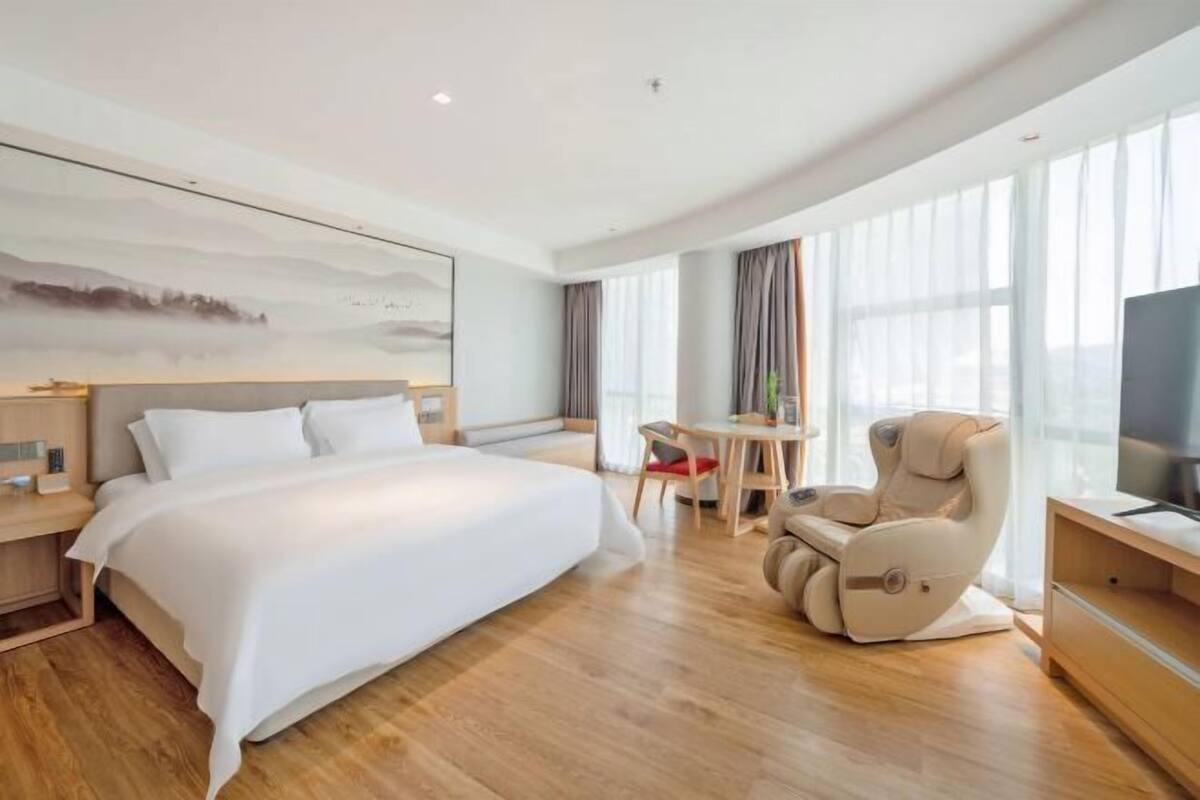 A modern hotel room with a large bed, a massage chair, a TV, and a dining area.