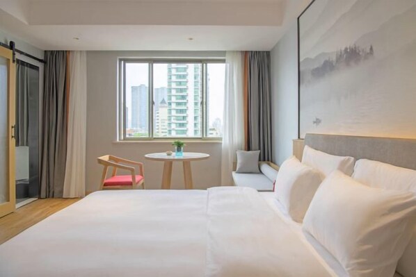 A hotel room with a large bed, a small table with a chair, a window with a view of buildings, and a painting on the wall.