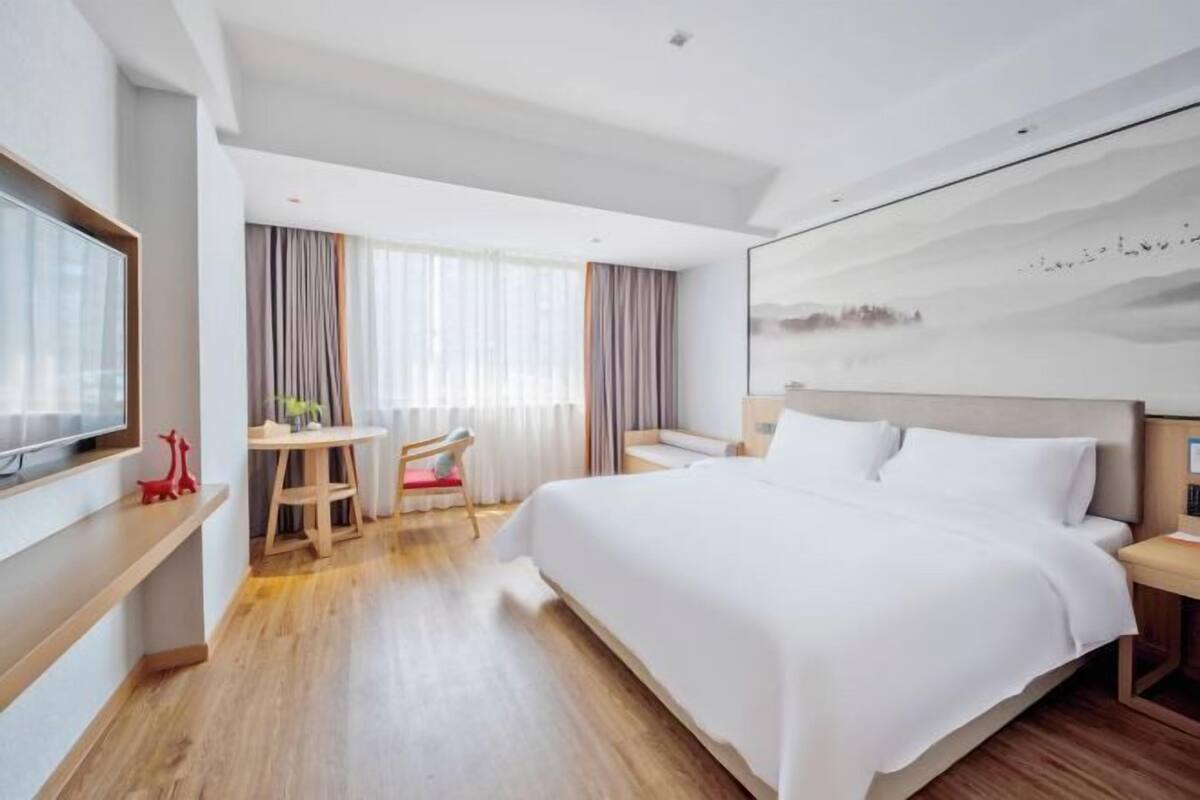 A modern hotel room with a large bed, a desk with a chair, and a view of the cityscape through the window.