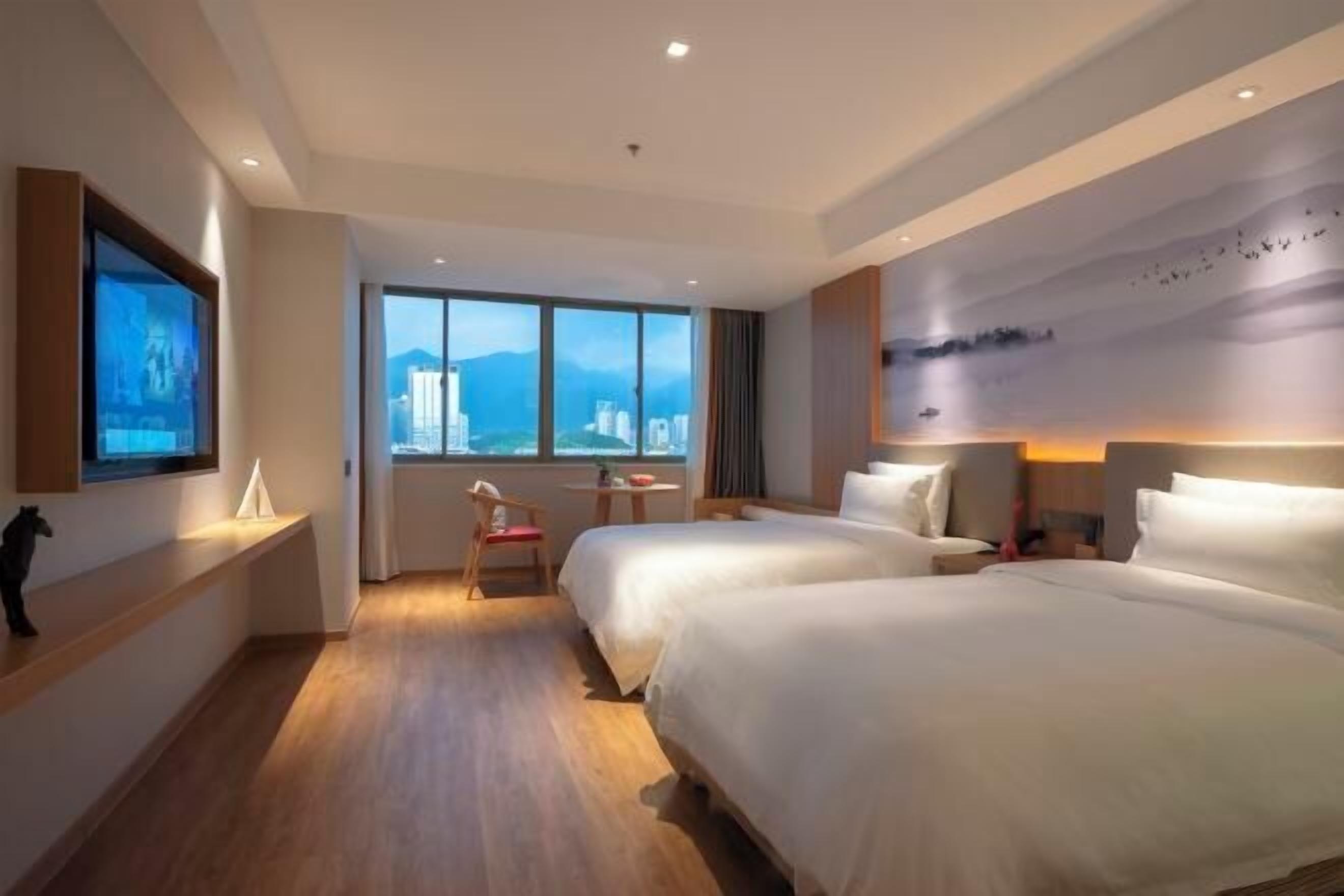 A hotel room with two beds, a large window with a city view, a small table with a chair, and a wall-mounted TV.