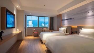 A hotel room with two beds, a large window with a city view, a small table with a chair, and a wall-mounted TV.