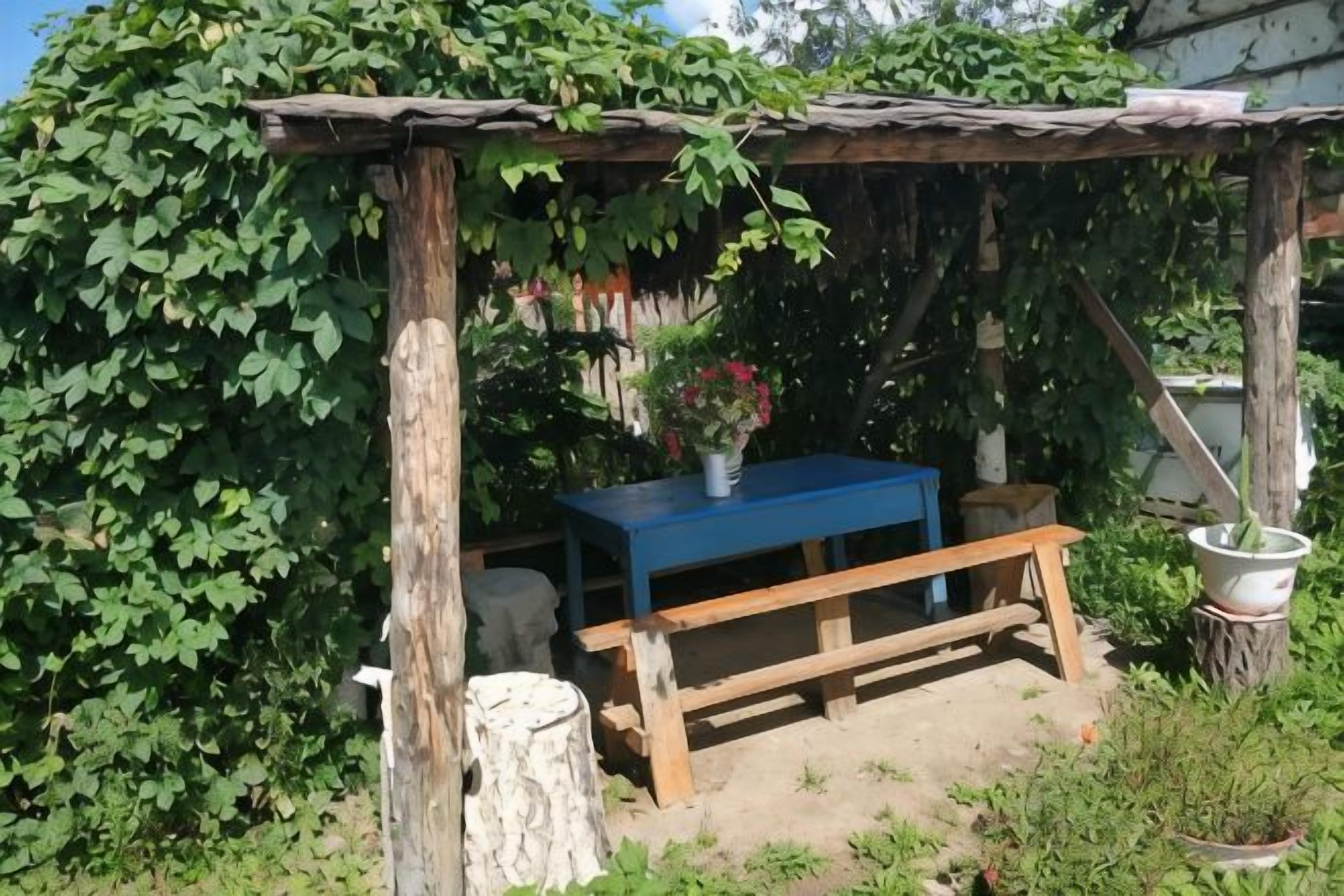 A rustic outdoor seating area with a wooden bench and a table, surrounded by greenery and a thatched roof structure.