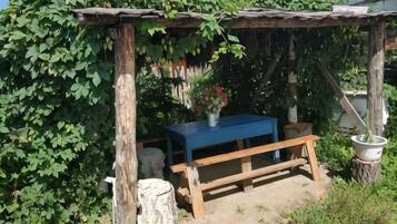 A rustic outdoor seating area with a wooden bench and a table, surrounded by greenery and a thatched roof structure.