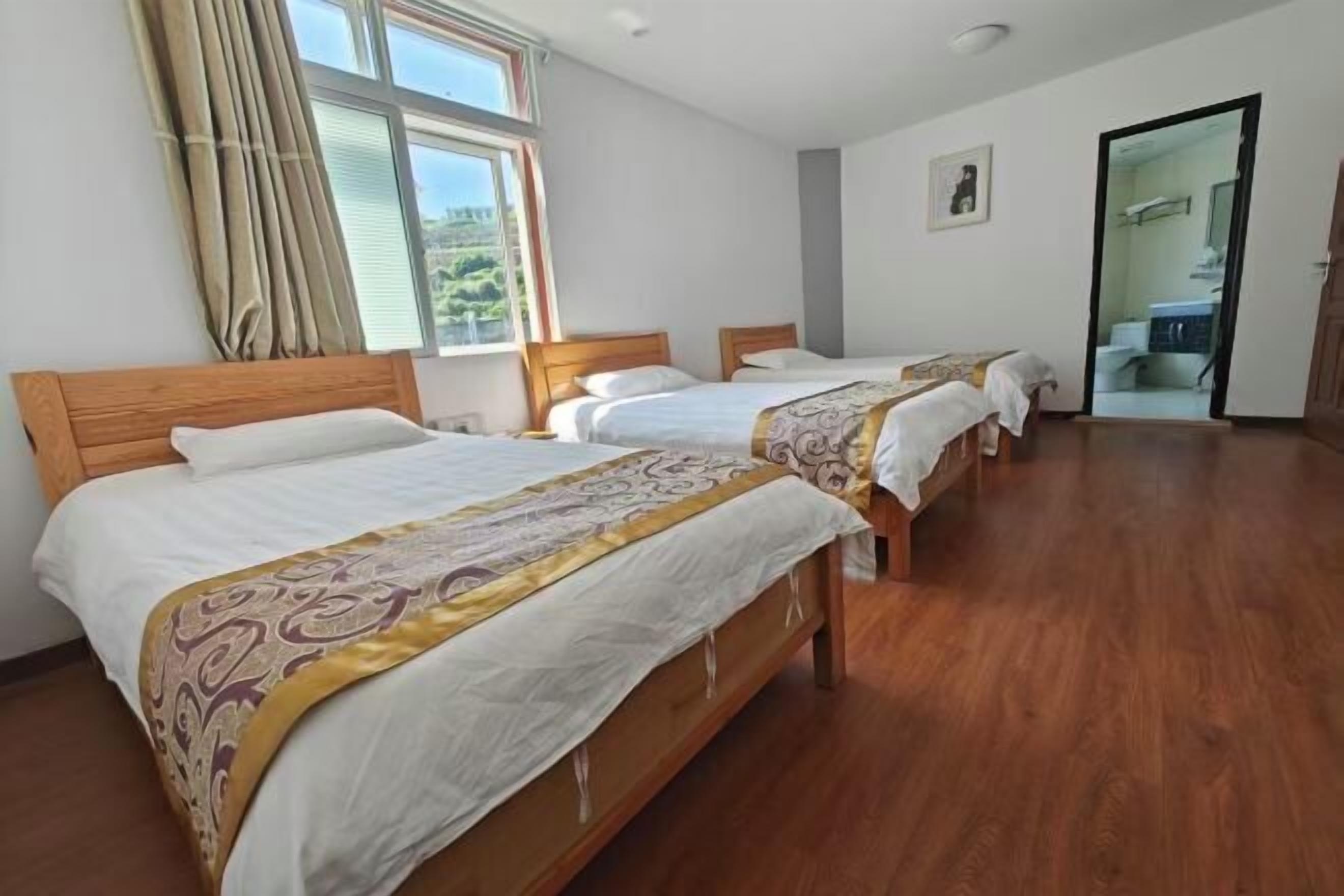 A hotel room with three wooden beds, a large window with curtains, and a door leading to a bathroom.
