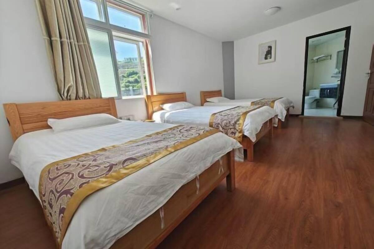 Een hotelkamer met drie houten bedden, een groot raam met gordijnen en een deur naar een badkamer.