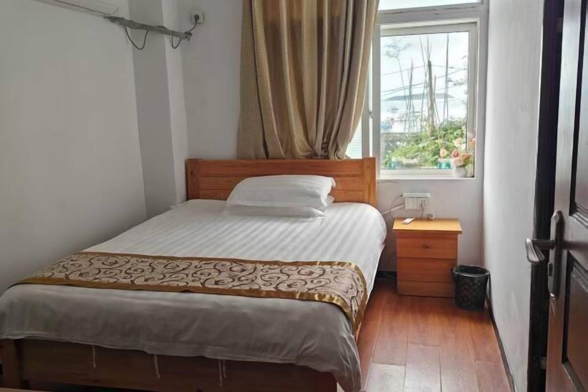 Een enkel bed met een houten hoofdbord, een nachtkastje met een lamp, een raam met gordijnen en uitzicht op groen buiten.