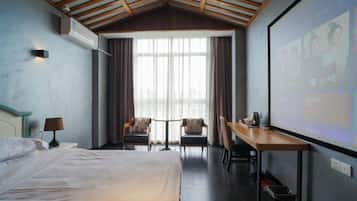 Habitación de hotel con cama, escritorio, dos sillas, pantalla para proyector y un ventanal con cortinas.