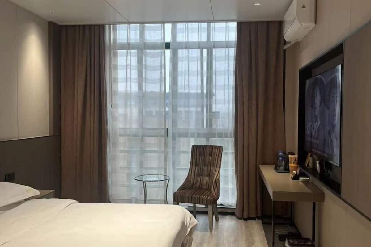 Hotellihuone, jossa on sänky, tuoli, pieni pöytä, televisio ja suuri ikkuna, jossa on verhot.