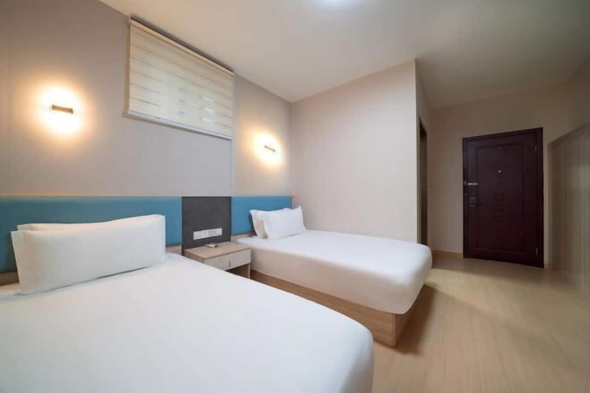A hotel room with two single beds, a bedside table with a lamp, a window with blinds, and a door.