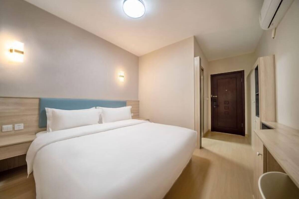 A hotel room with a large bed, a wooden headboard, a bedside table, a nightstand, a wall-mounted light, an air conditioning unit, and a door leading to another room.