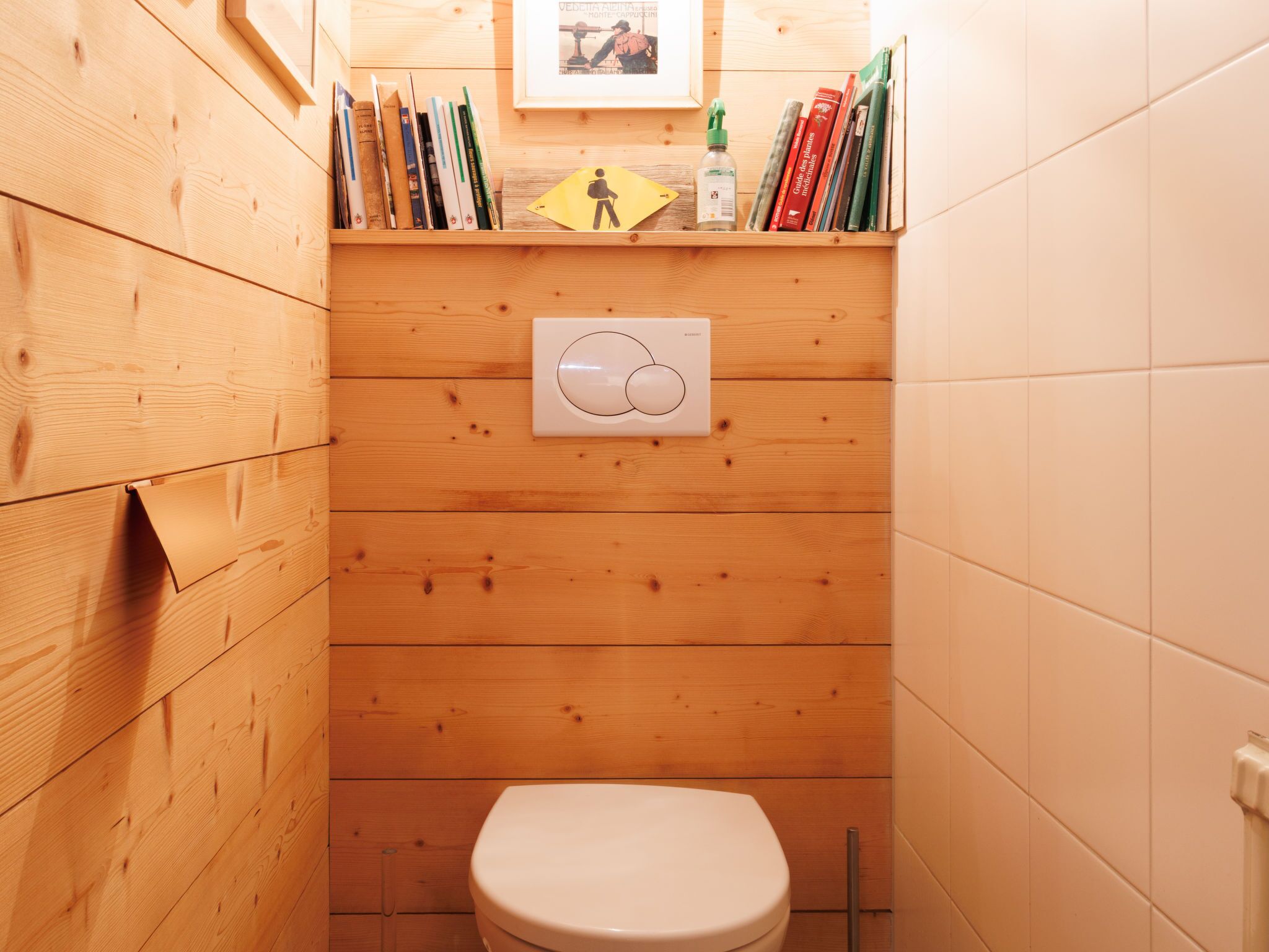 A small, wooden-paneled bathroom with a toilet, a hand dryer, and a shelf with books and a framed picture.