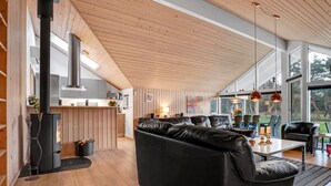 A modern living room with a black leather sofa, wooden flooring, a kitchen area with a stove, and large windows offering a view of the outdoors.