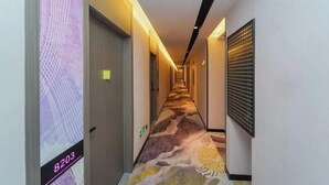 Een hotelgang met een tapijtvloer, deuren aan de linkerkant en een ventilatie rooster aan de rechterkant.