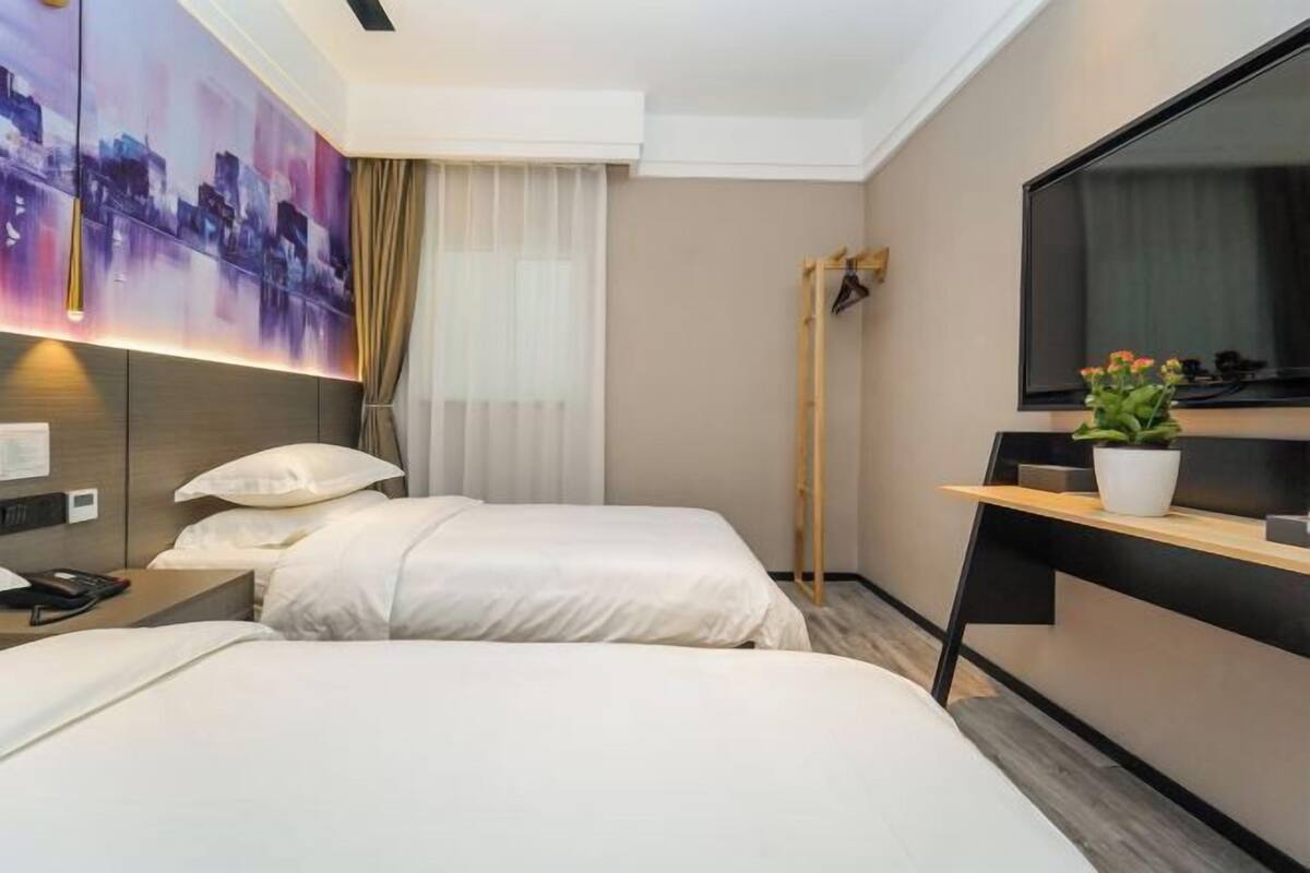 A hotel room with two beds, a large TV, a painting on the wall, and a plant on a side table.