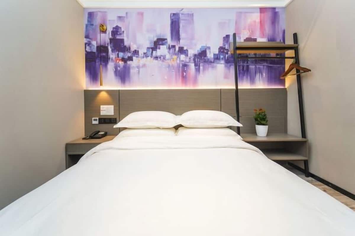 A hotel room with a bed, a nightstand with a phone, a wall-mounted shelf with a plant, and a cityscape painting above the bed.