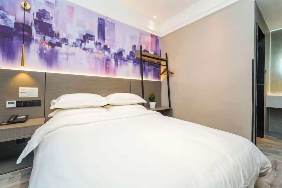 A modern hotel room with a large bed, a cityscape painting on the wall, a small plant on the nightstand, and a desk with a phone and a lamp.