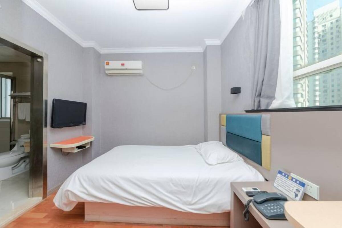 A hotel room with a bed, a TV, an air conditioning unit, a bedside table with a phone, and a window with a city view.