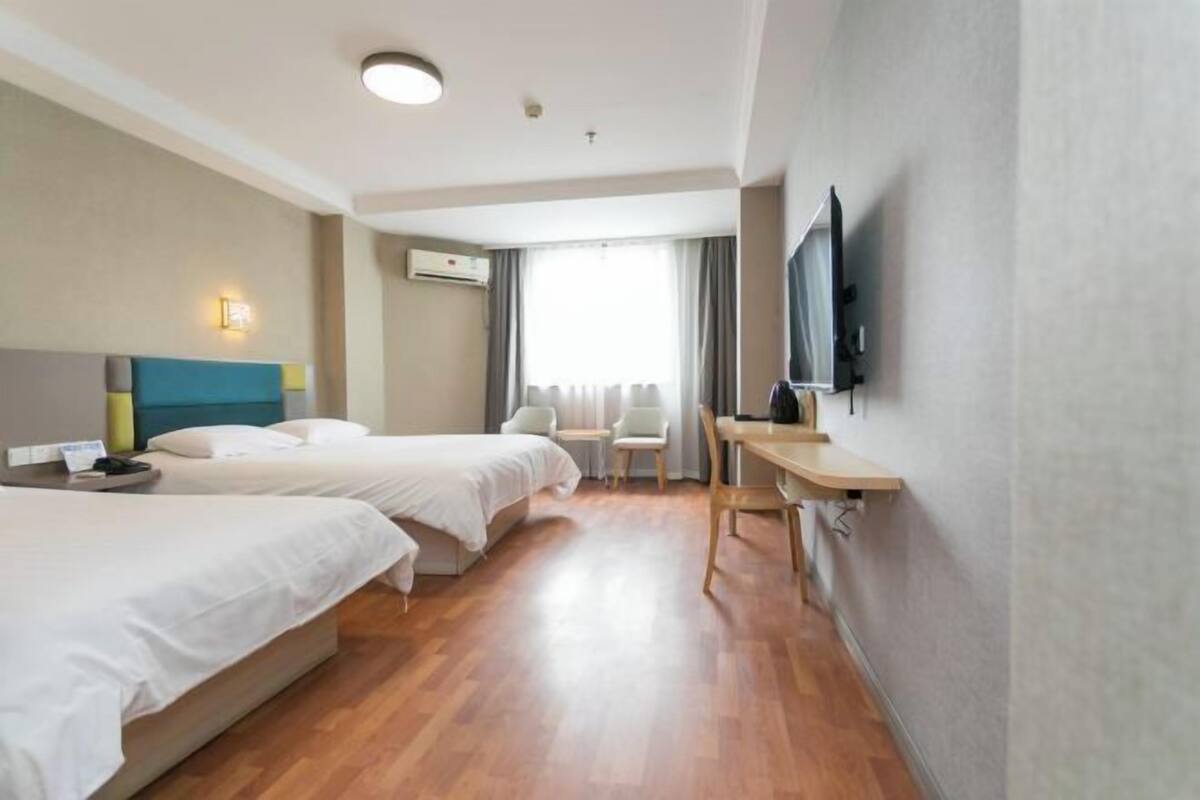 A hotel room with two beds, a desk, a chair, a flat-screen TV, and a window with curtains.