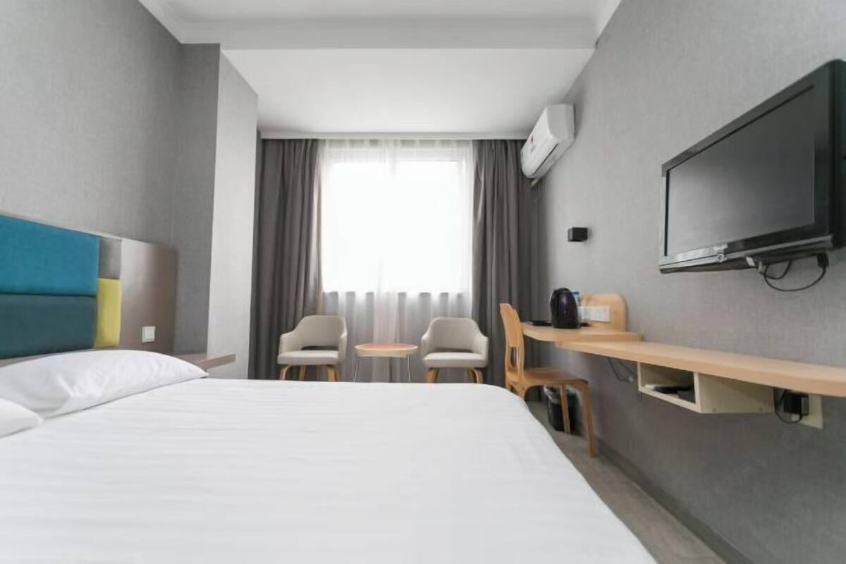 A hotel room with a bed, a TV, a desk with a chair, and a window with curtains.