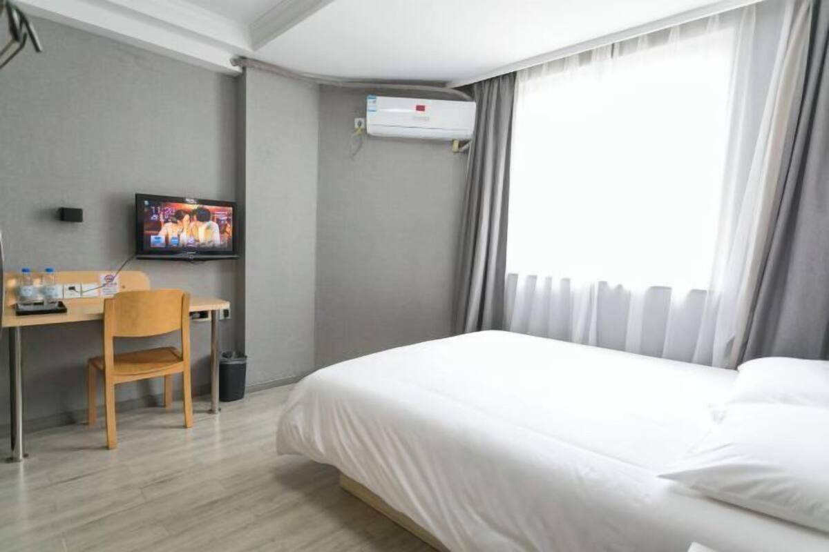 A hotel room with a bed, a desk with a chair, a television, and an air conditioning unit.