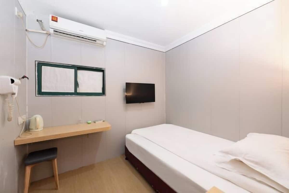 A small, clean room with a bed, a desk, a chair, a hairdryer, and a TV mounted on the wall.