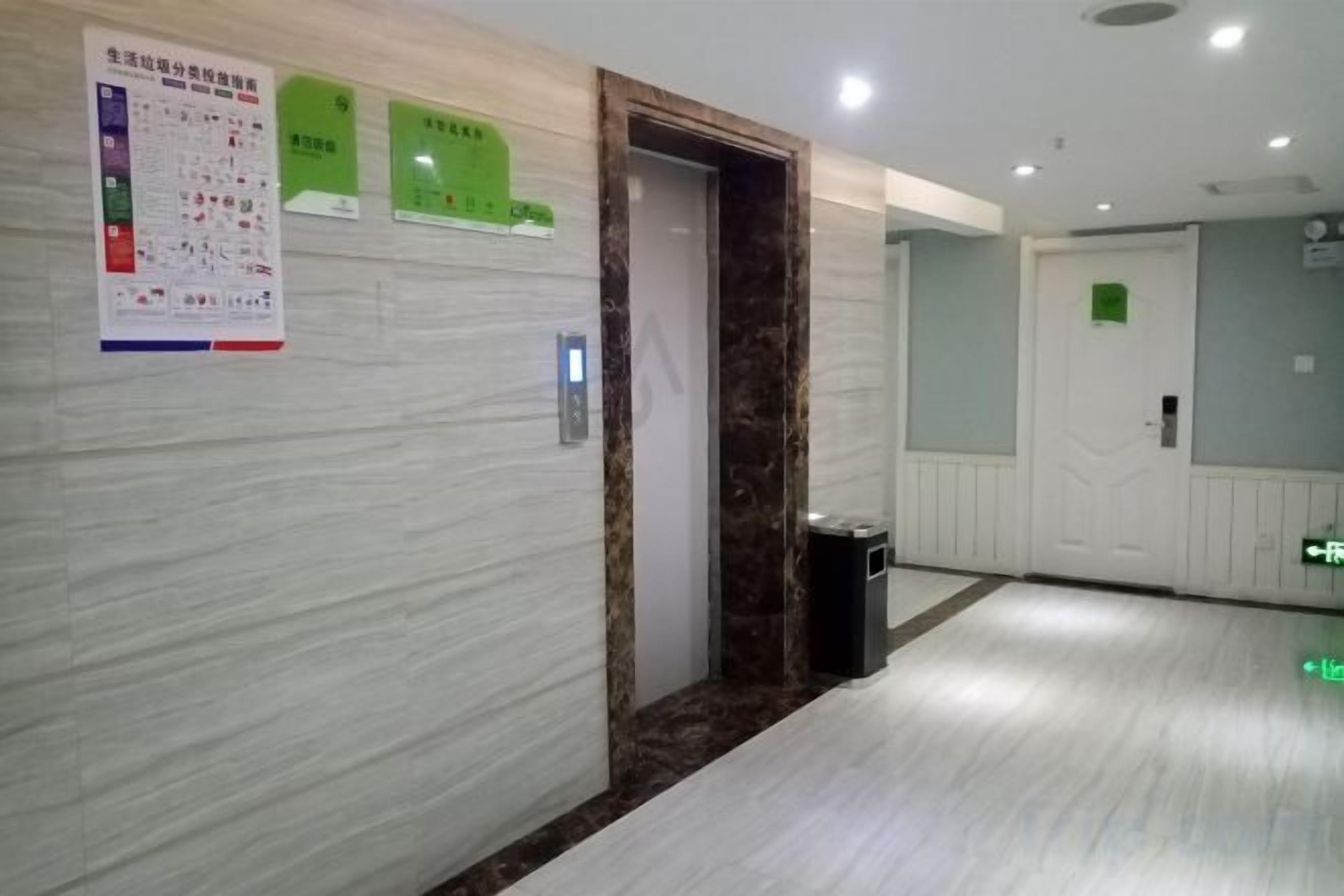 A hotel corridor with a marble wall, a door with a green sign, and a trash bin.