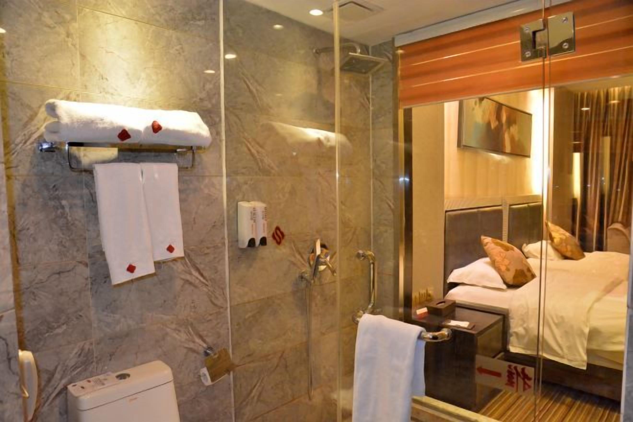 A hotel room with a glass shower enclosure, a bed with pillows, and a bedside table.