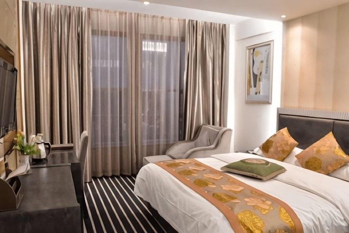 Habitación de hotel con cama, cortinas, cabecero, un cuadro y escritorio con silla.