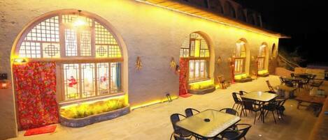 Un área de comedor al aire libre con mesas y sillas, amplias ventanas de arco y fachada de edificio.