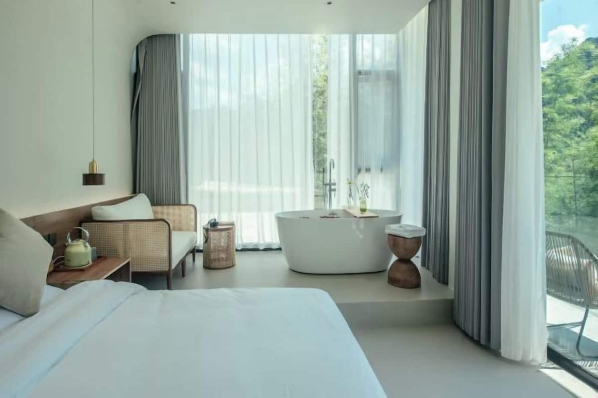 A modern bedroom with a large bed, a sofa, a bathtub, and a view of greenery outside.