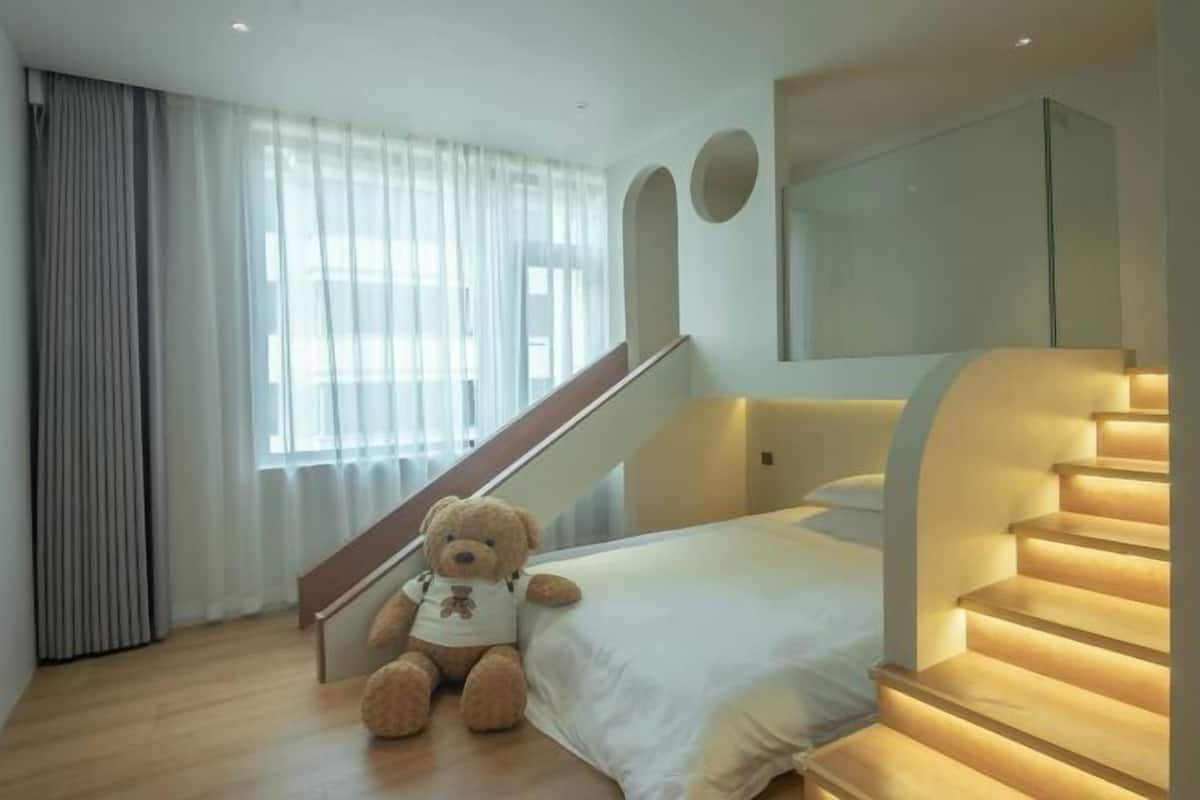 A bedroom with a bed, a teddy bear, a staircase, and a window with curtains.