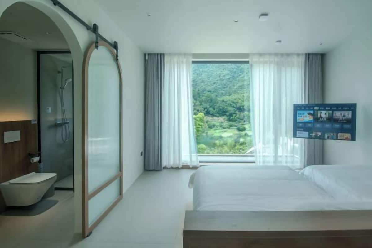 A hotel room with a bed, a shower, and a large window with curtains.
