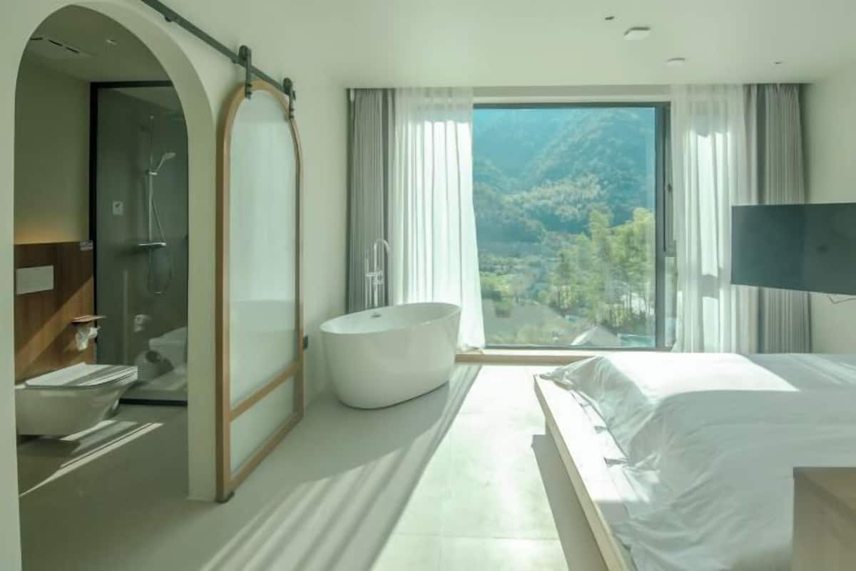 A modern hotel room with a large bed, a bathtub, a vanity with a mirror, a shower, and a large window with a mountain view.