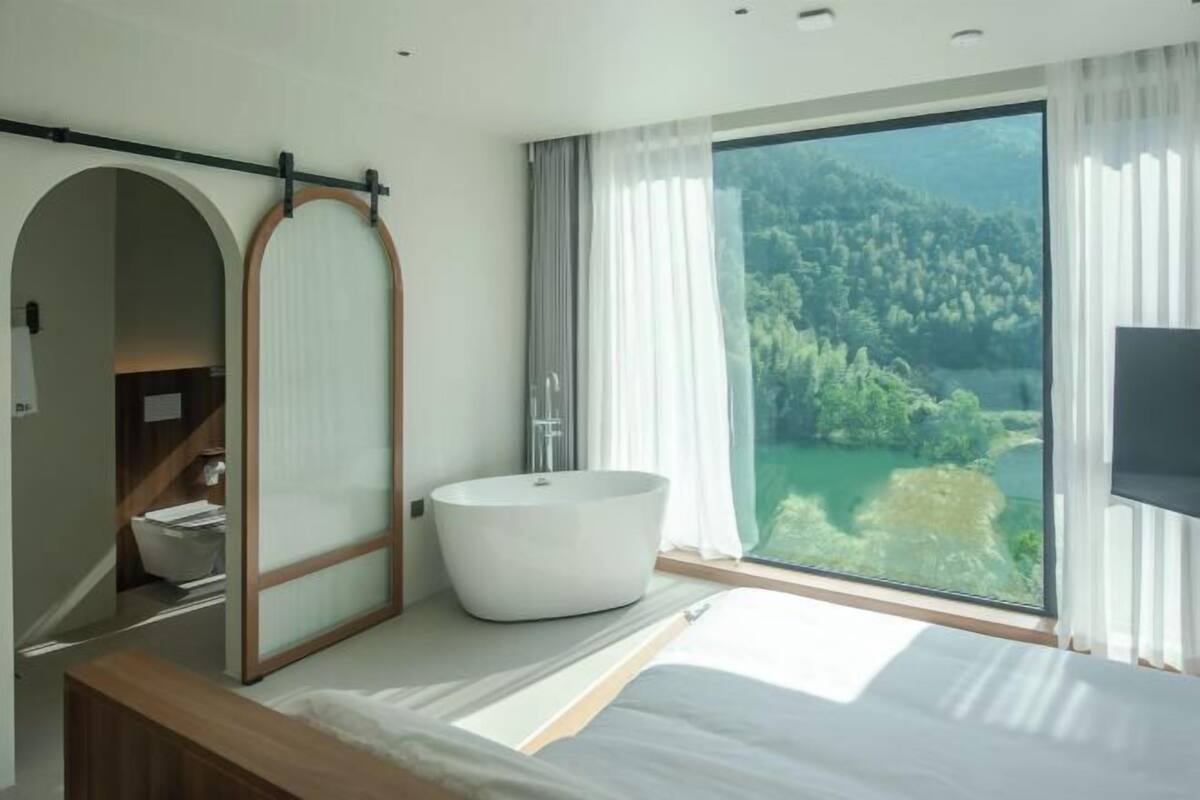 A modern bedroom with a large window, a bathtub, and a wooden vanity.