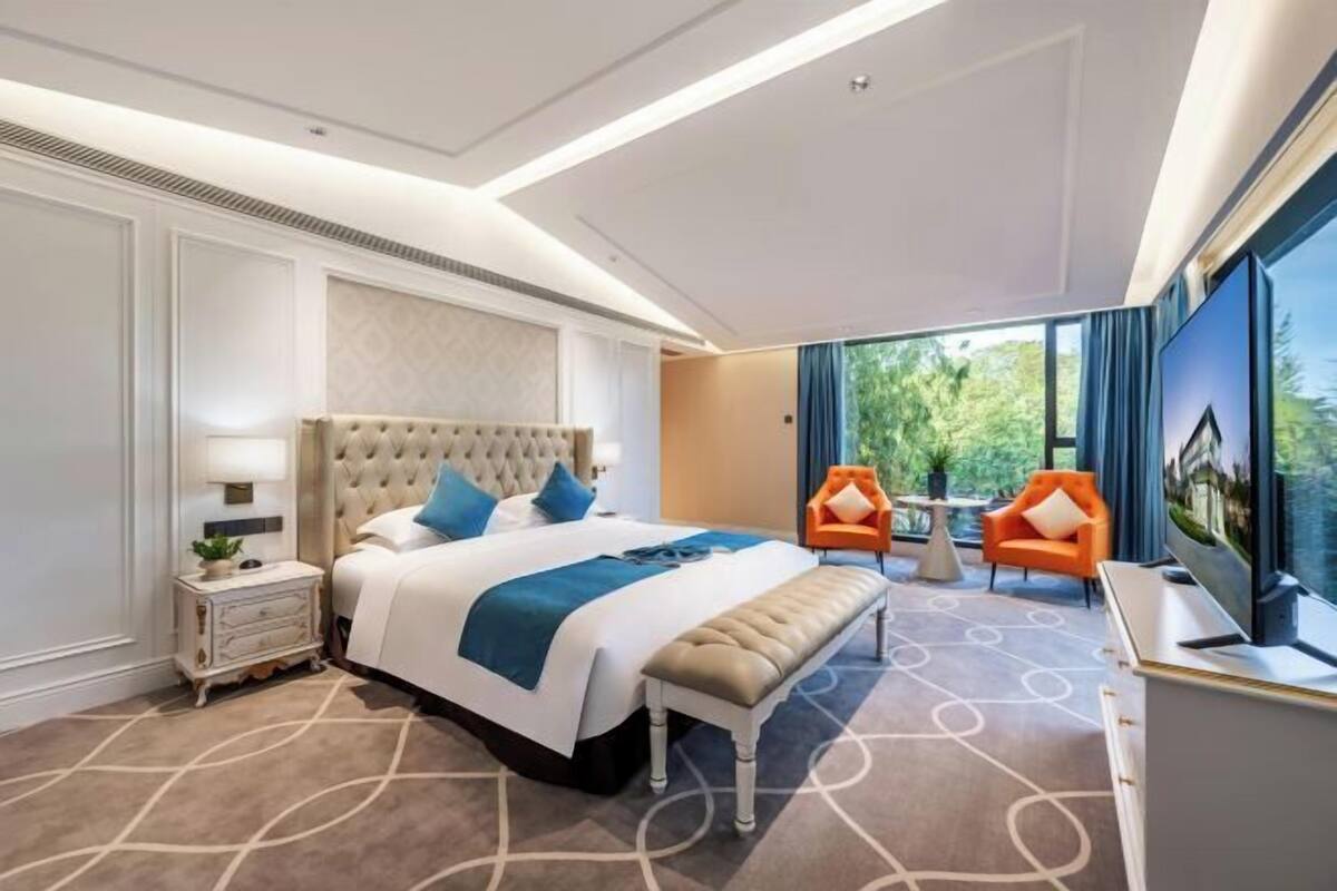A hotel room with a large bed, a bench, two chairs, a TV, and a view of greenery outside.