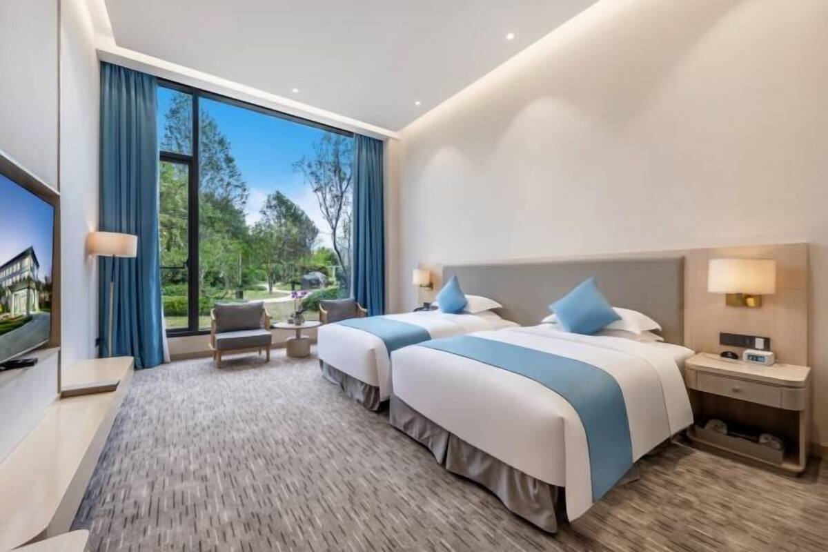 A hotel room with two beds, a TV, a desk, and a large window with curtains.