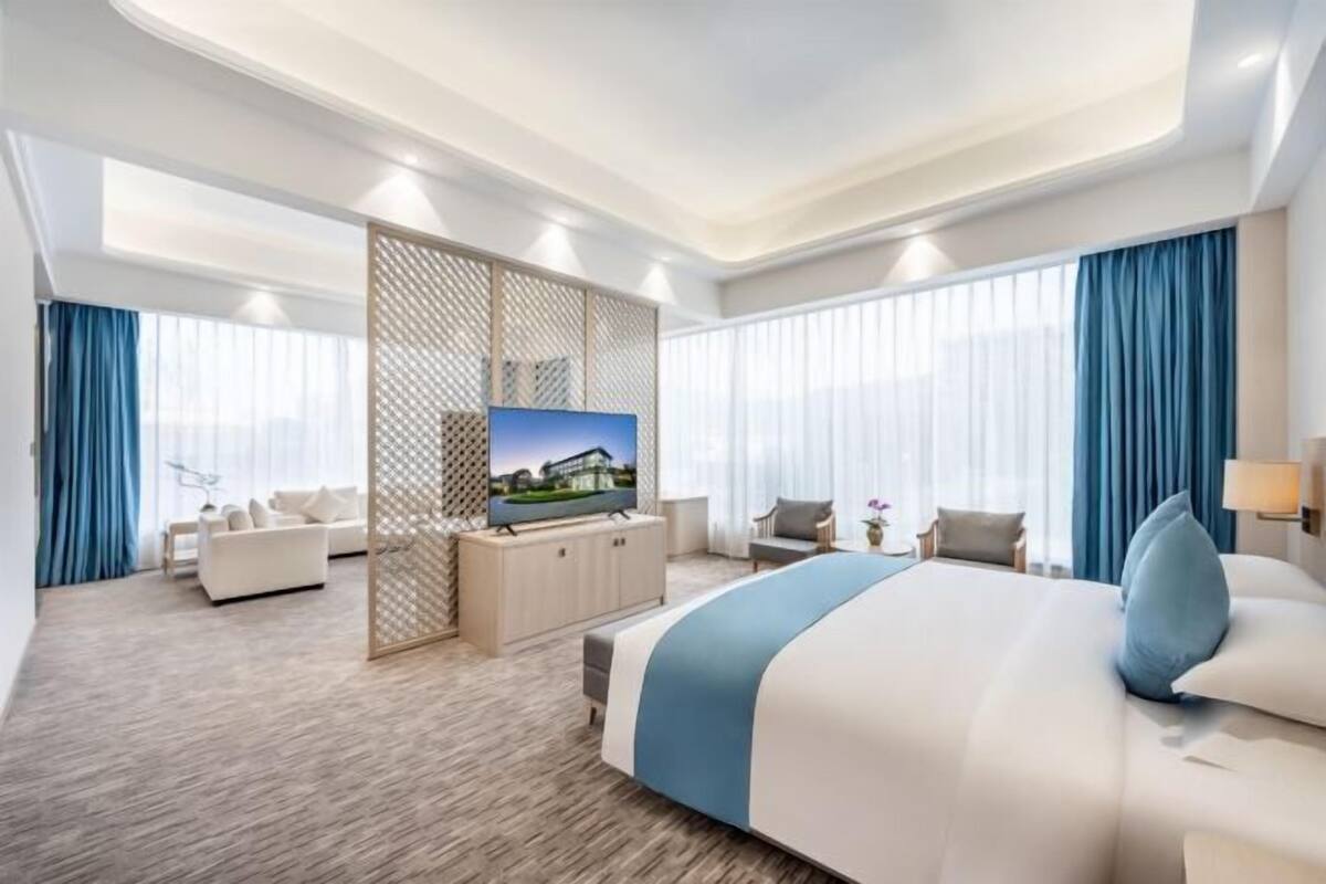 A modern hotel room with a large bed, a TV, a seating area with a sofa and chairs, and a large window with curtains.