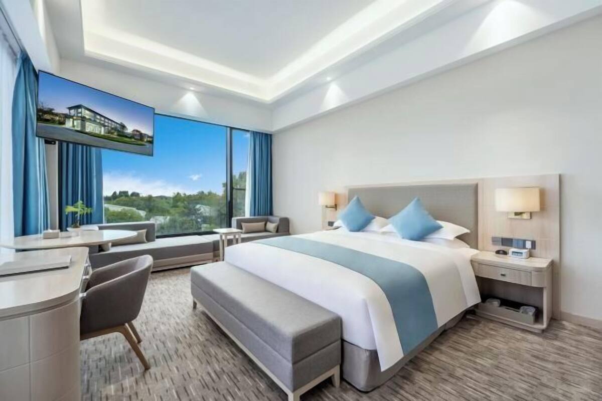 A modern hotel room with a large bed, a desk, a bench, and a view of a building through the window.