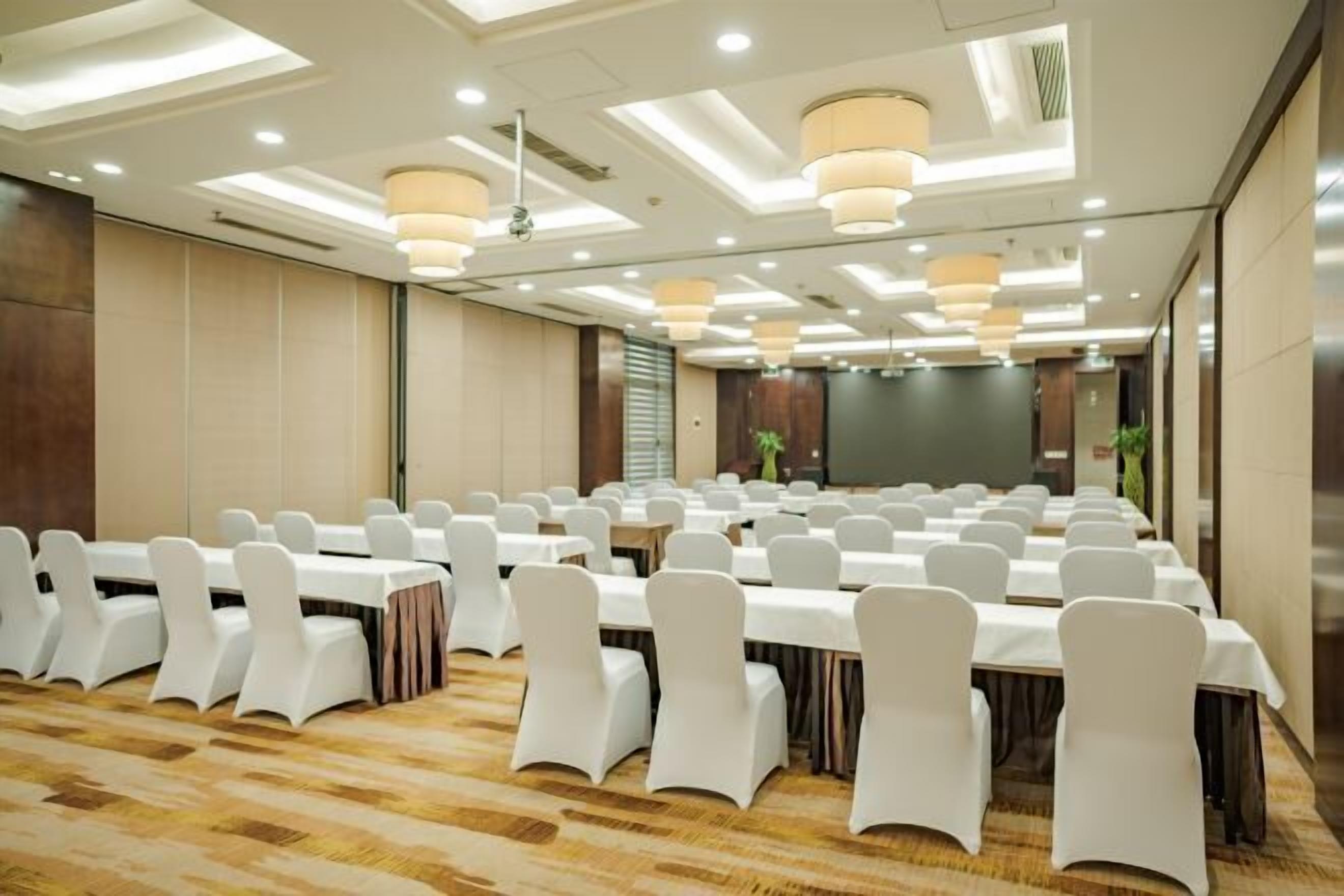 Un salón de conferencias con filas de mesas y sillas, iluminación empotrada y una pantalla de proyección.