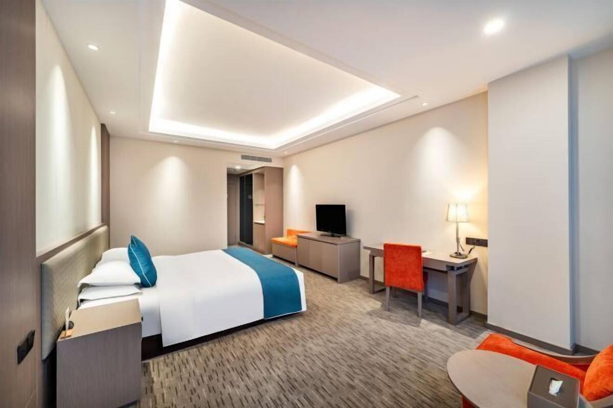 A modern hotel room with a bed, desk, chair, TV, and a small table with a lamp.