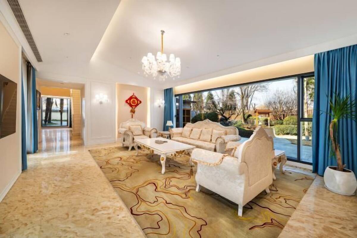 A spacious living room with a large window, a chandelier, and a decorative wall piece.