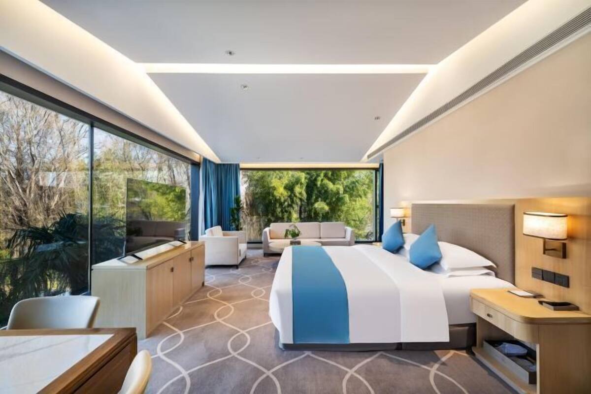 A modern hotel room with a large bed, a desk, a chair, and a view of greenery outside.