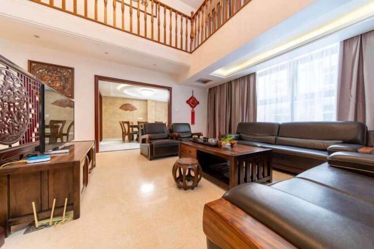 Un salón amplio con techo alto, una escalera de madera, un conjunto de sofás, una mesa de centro y un televisor sobre un mueble de madera.