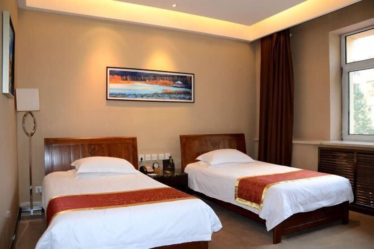A hotel room with two single beds, a nightstand with a lamp, a painting on the wall, and a window with curtains.