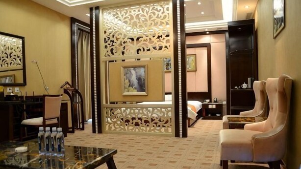 A hotel room with a bed, a sitting area with a chair, a table with water bottles, a decorative screen, and artwork on the walls. - Mohe International Conference Center (Mohe)