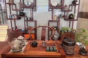 Un juego de té tradicional sobre una mesa de madera, con una silla, rodeado de estantes con cerámica variada y plantas.