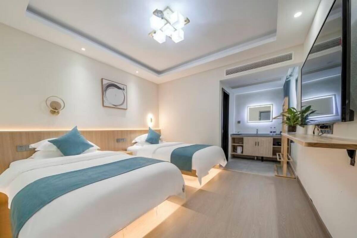 A modern hotel room with two beds, a small kitchenette, and a bathroom visible through an open door.