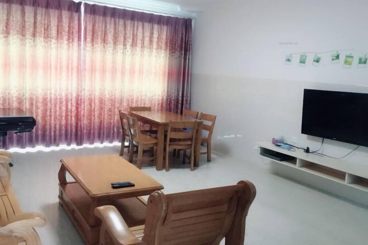 Uma sala de jantar com uma mesa, cadeiras, uma televisão em um suporte e um piano ao fundo.