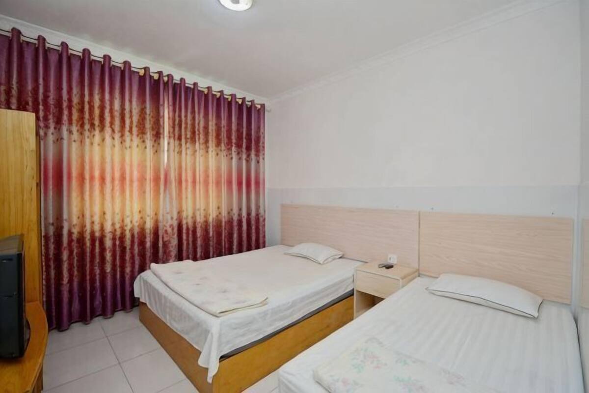 Quarto com duas camas, uma mesa de cabeceira, um guarda-roupa de madeira e uma cortina.