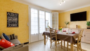 Un coin repas avec une table dressée pour quatre personnes, un canapé avec des coussins, un téléviseur fixé au mur et une porte-fenêtre coulissante donnant sur un espace extérieur.